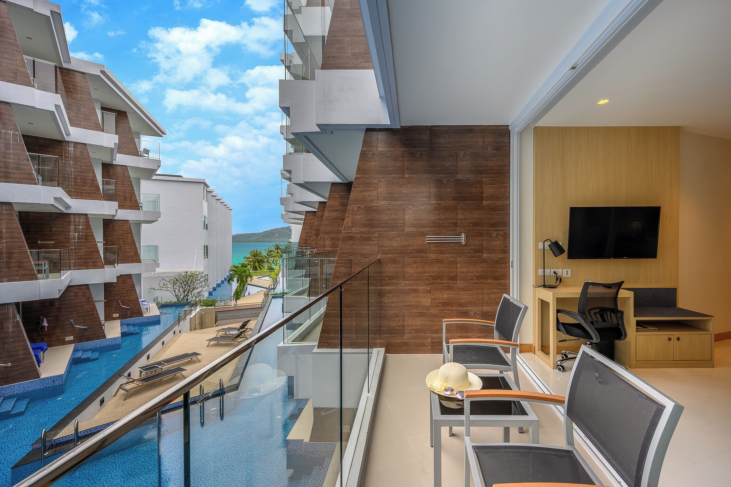 A modern hotel room with a glass balcony overlooking a swimming pool, a wooden wall, and a TV in the corner.