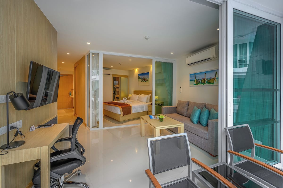 A modern hotel room with a bed, a sofa, a desk with a chair, a TV, and a large window.
