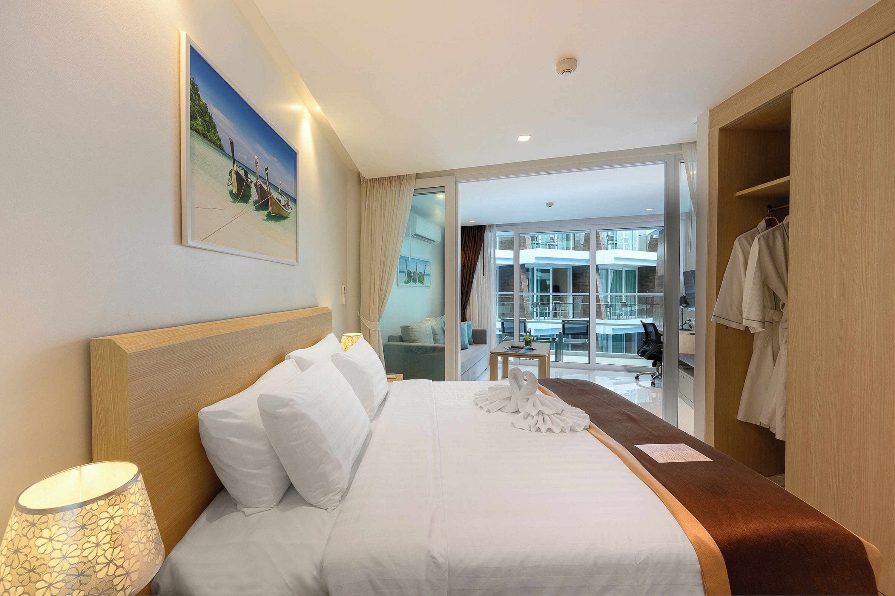 Habitación de hotel con una cama bien hecha, una lámpara de noche, un cuadro con una escena de playa y vistas del exterior del hotel a través de un ventanal.