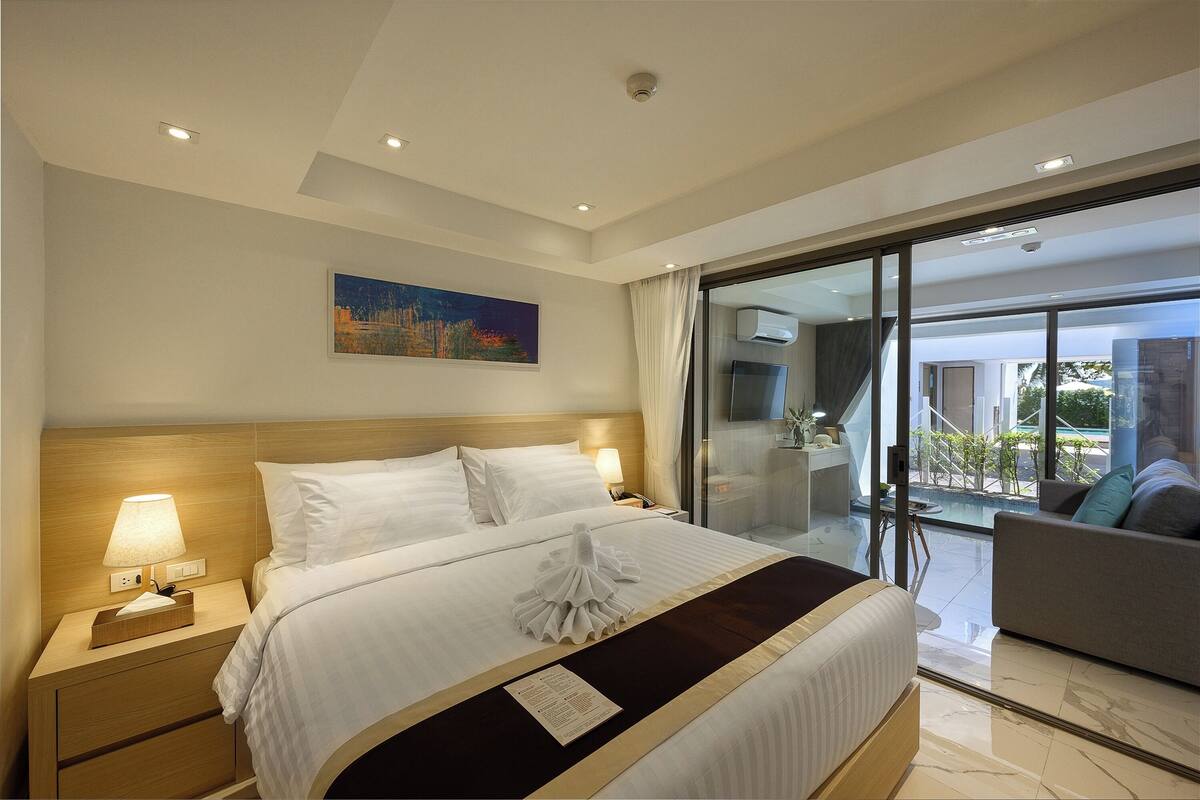 A hotel room with a large bed, a bedside table with a lamp, a painting on the wall, and a view of the outdoors through a glass door.