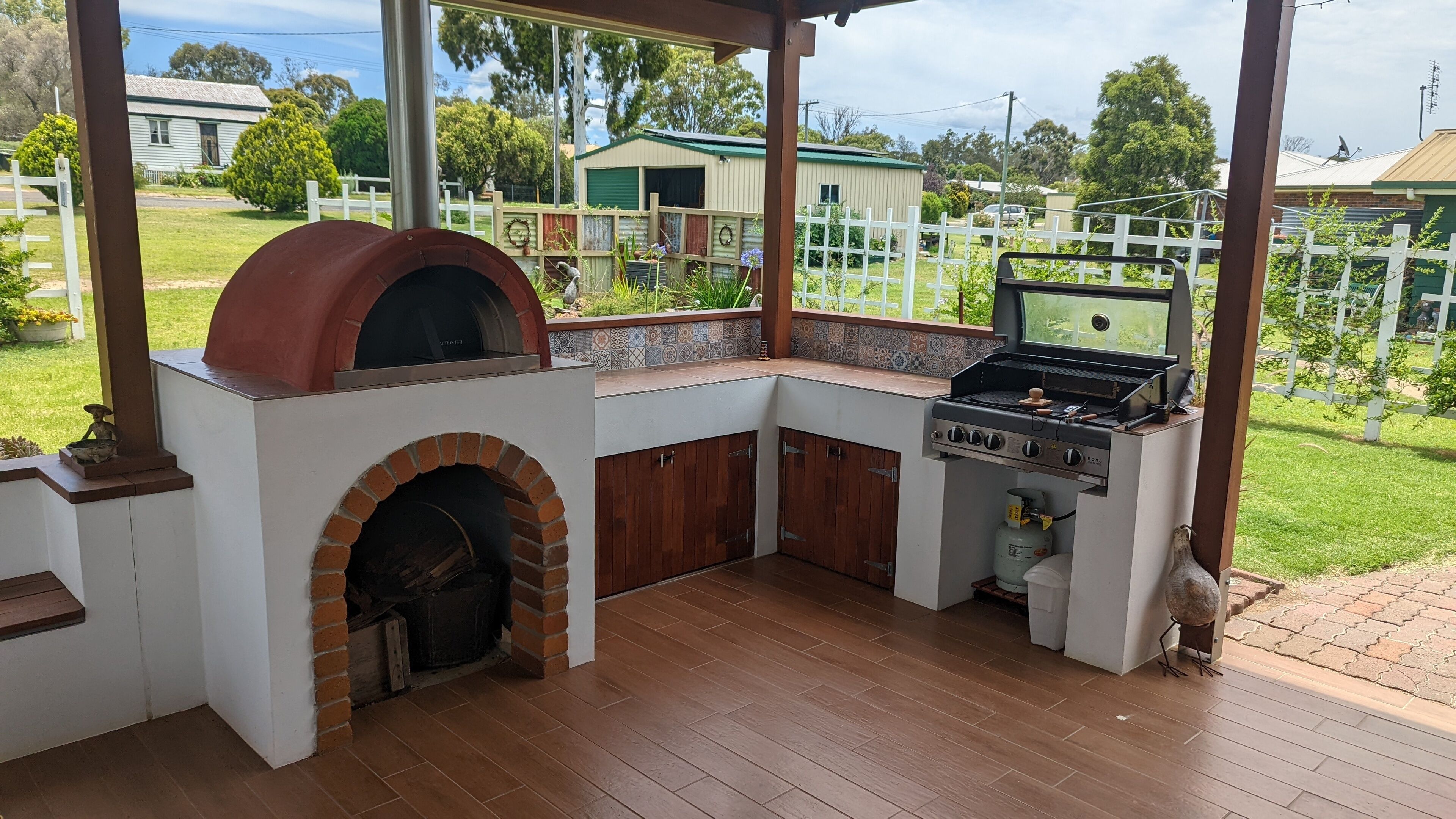 Eine Außenküche mit Backofen aus Ziegeln und Gasgrill, umgeben von einer Holzterrasse und einem weißen Zaun.