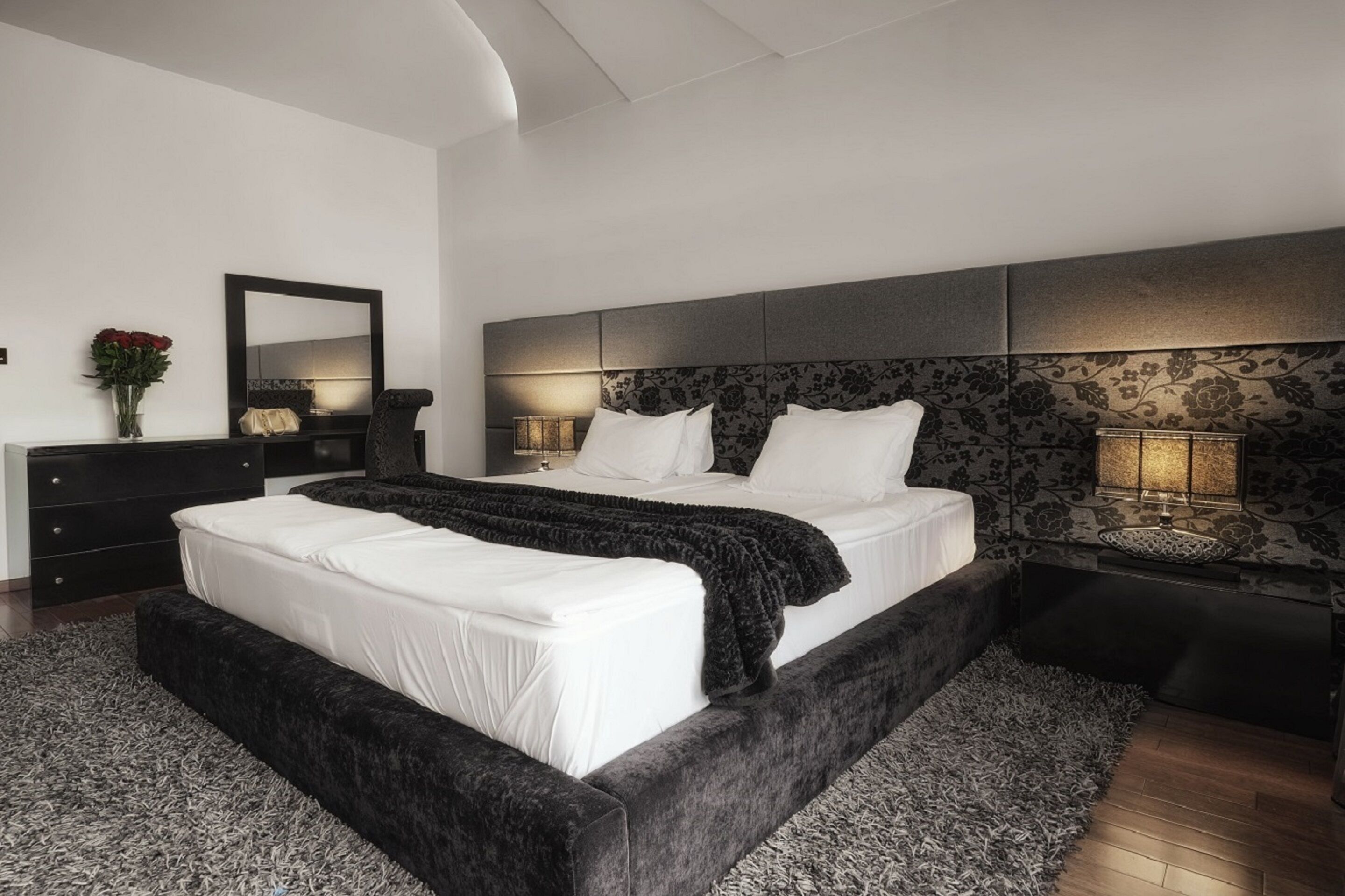 Ein modernes Schlafzimmer mit einem großen Bett, einem dunklen Kopfteil, Nachttischlampen und einem Spiegel.