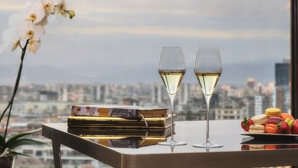 A table with two glasses of white wine, a stack of books, and a plate of colorful macarons, set against a cityscape view. - Hotel Emirates (Sofia)