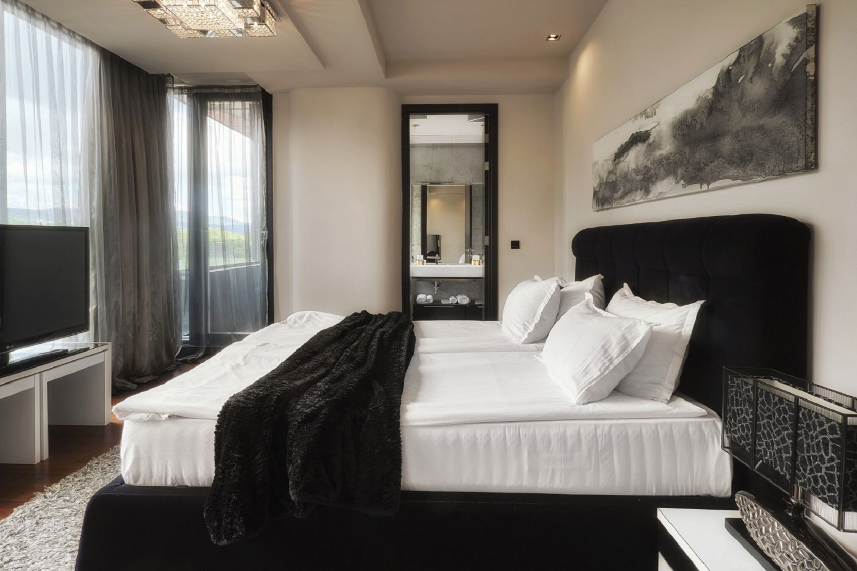 A hotel room with two beds, a large flat-screen TV, a painting on the wall, and a view of the outdoors through the window.