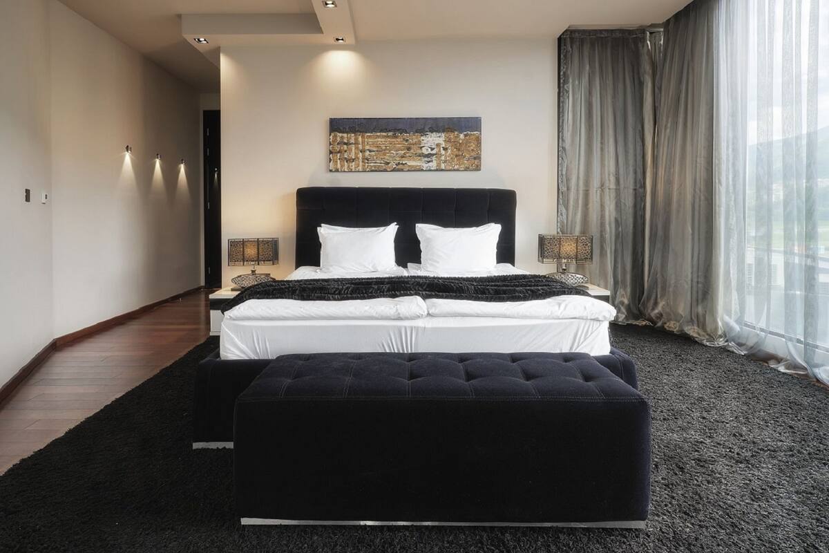 A modern bedroom with a large bed, a black ottoman, and a painting on the wall.