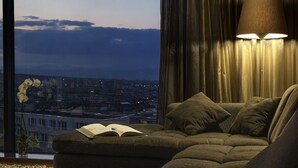 A cozy living room with a sofa, a lamp, and a view of a cityscape through a window. - Hotel Emirates (Sofia)
