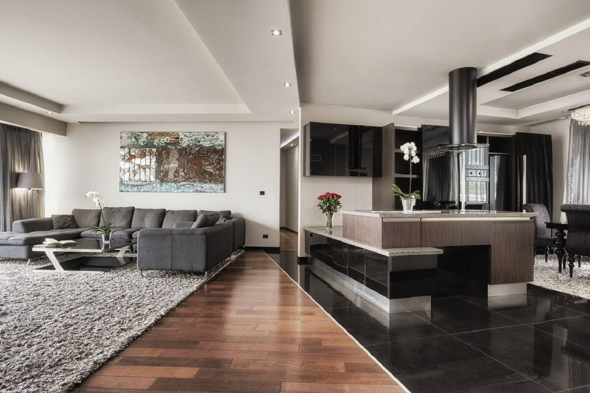 A modern living room with a sofa, coffee table, and a rug. There is a kitchen area with a stove and cabinets.