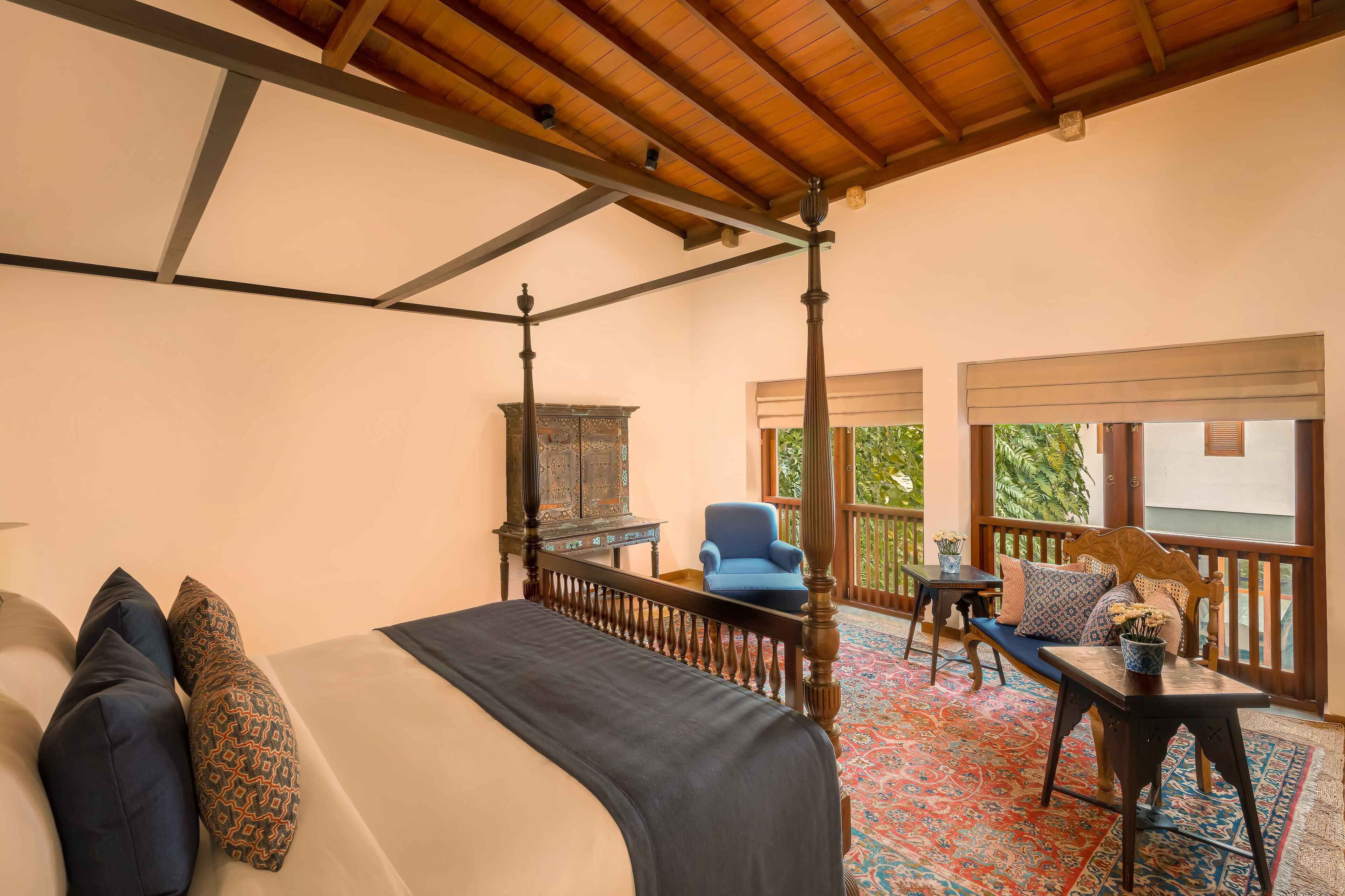 A four-poster bed with a dark bedspread and patterned pillows, a wooden ceiling, a small table with a vase of flowers, and a blue chair.
