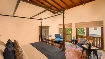 A four-poster bed with a dark bedspread and patterned pillows, a wooden ceiling, a small table with a vase of flowers, and a blue chair.