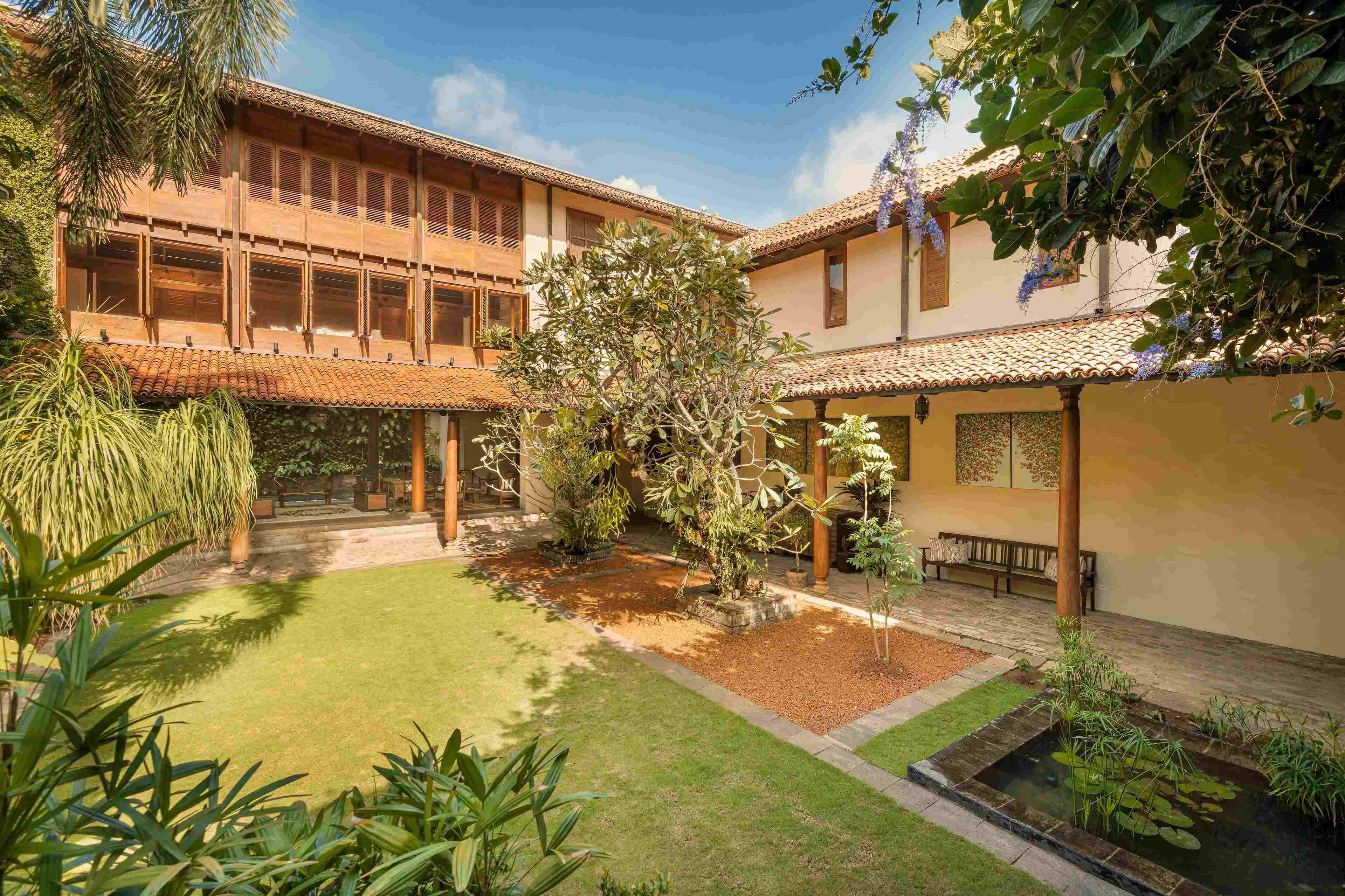 A courtyard with a building featuring a tiled roof and wooden balconies, surrounded by lush greenery and a small water feature.