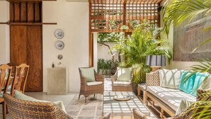 A cozy indoor space with wicker furniture, a wooden bench, and a sofa with cushions. There are potted plants and a wooden lattice structure.