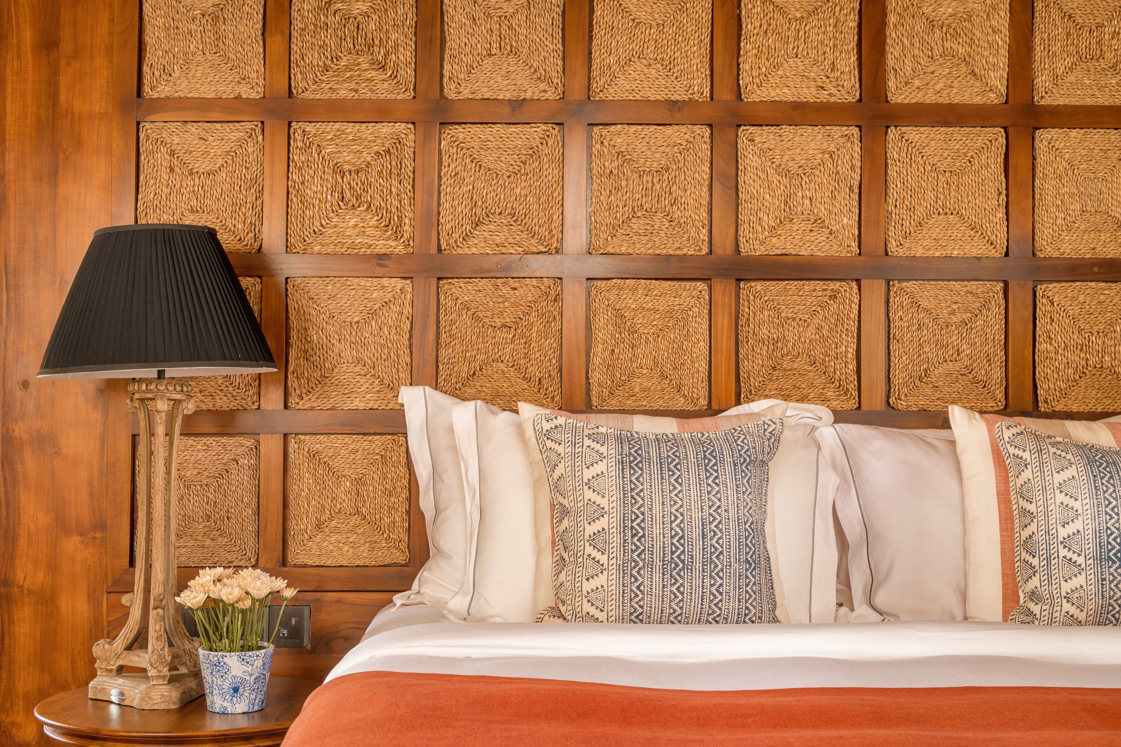 A neatly made bed with patterned pillows, a bedside lamp, and a vase of flowers on a wooden nightstand.