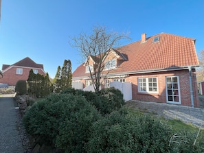 Uma casa residencial com telhado de telhas vermelhas, paredes de tijolo e um jardim bem cuidado, com sebes e uma árvore.