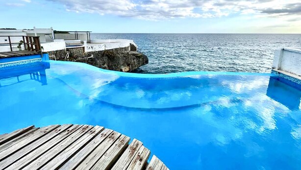 A pool with a wooden deck, a white building with a balcony, and a view of the ocean. - ON THE Cliff IN THE Caribbean SEA - Villa Ilaria (Boca Chica)