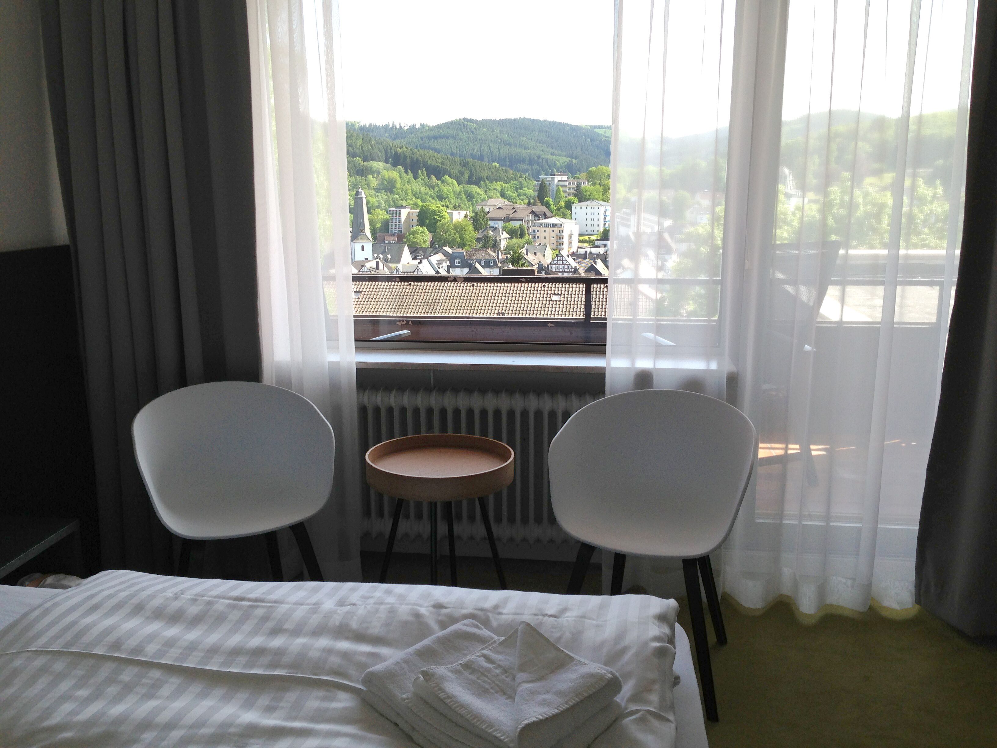 Habitació d'hotel amb un llit, dues cadires, una taula petita i vista a una vila i un bosc a través de cortines primes.