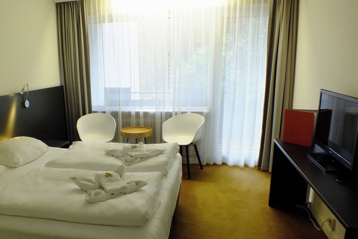 Habitación de hotel con cama, dos sillas, una mesita, televisor y ventana con cortinas.