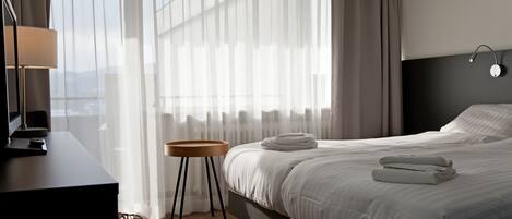 Habitación de hotel con cama, escritorio, lámpara y vistas al exterior a través de cortinas translúcidas.
