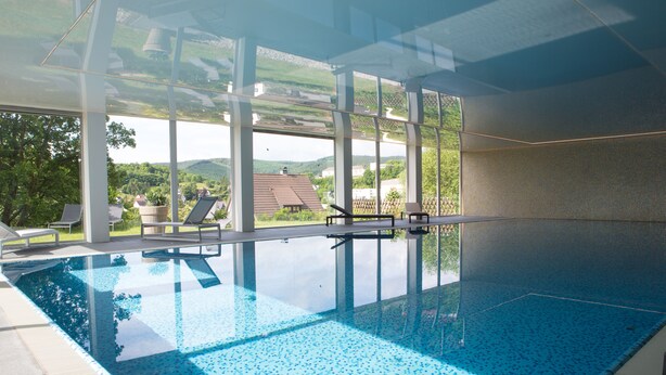An indoor swimming pool with clear glass walls, lounge chairs, and a view of the surrounding landscape. - Hotel Am Fang (Bad Laasphe)