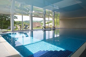 An indoor swimming pool with clear glass walls, lounge chairs, and a view of the surrounding landscape. - Hotel Am Fang (Bad Laasphe)