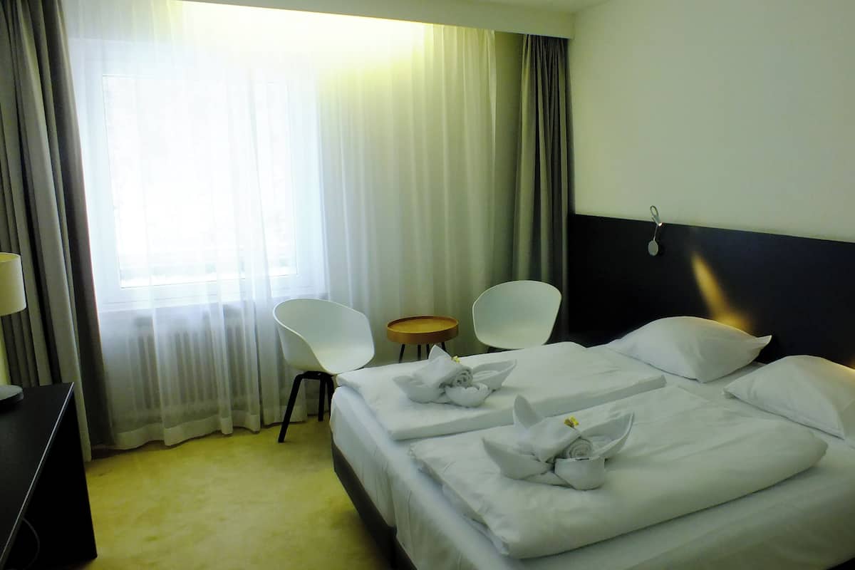 Habitación de hotel con dos camas, una mesita, dos sillas y una ventana con cortinas.