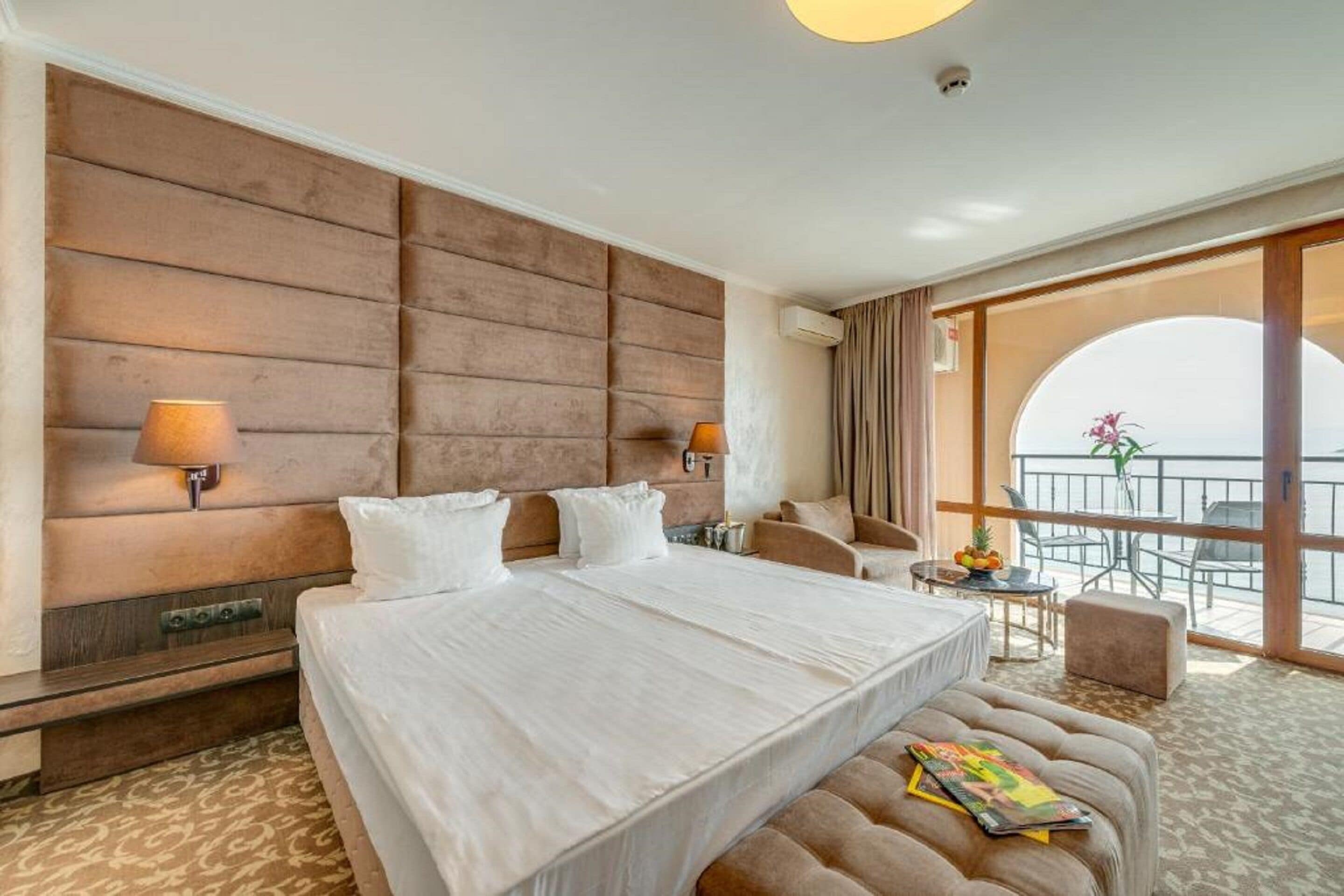 Hotelkamer met een groot bed, twee bedlampen, een bank, een kleine tafel met fruit en een balkon met uitzicht.