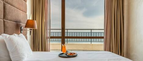 A hotel room with a bed, a bedside table with a lamp, a tray with food and a drink, and a view of the sea through large windows.
