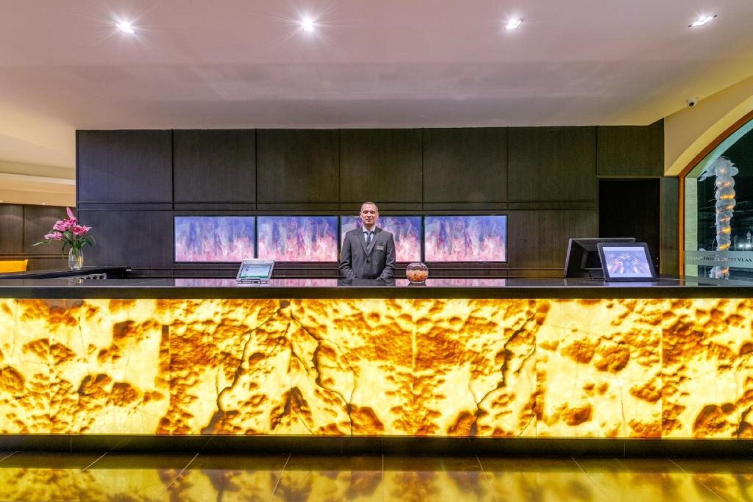 A reception desk with a large screen displaying a fire image, a man standing behind it, and a decorative vase with flowers to the left.