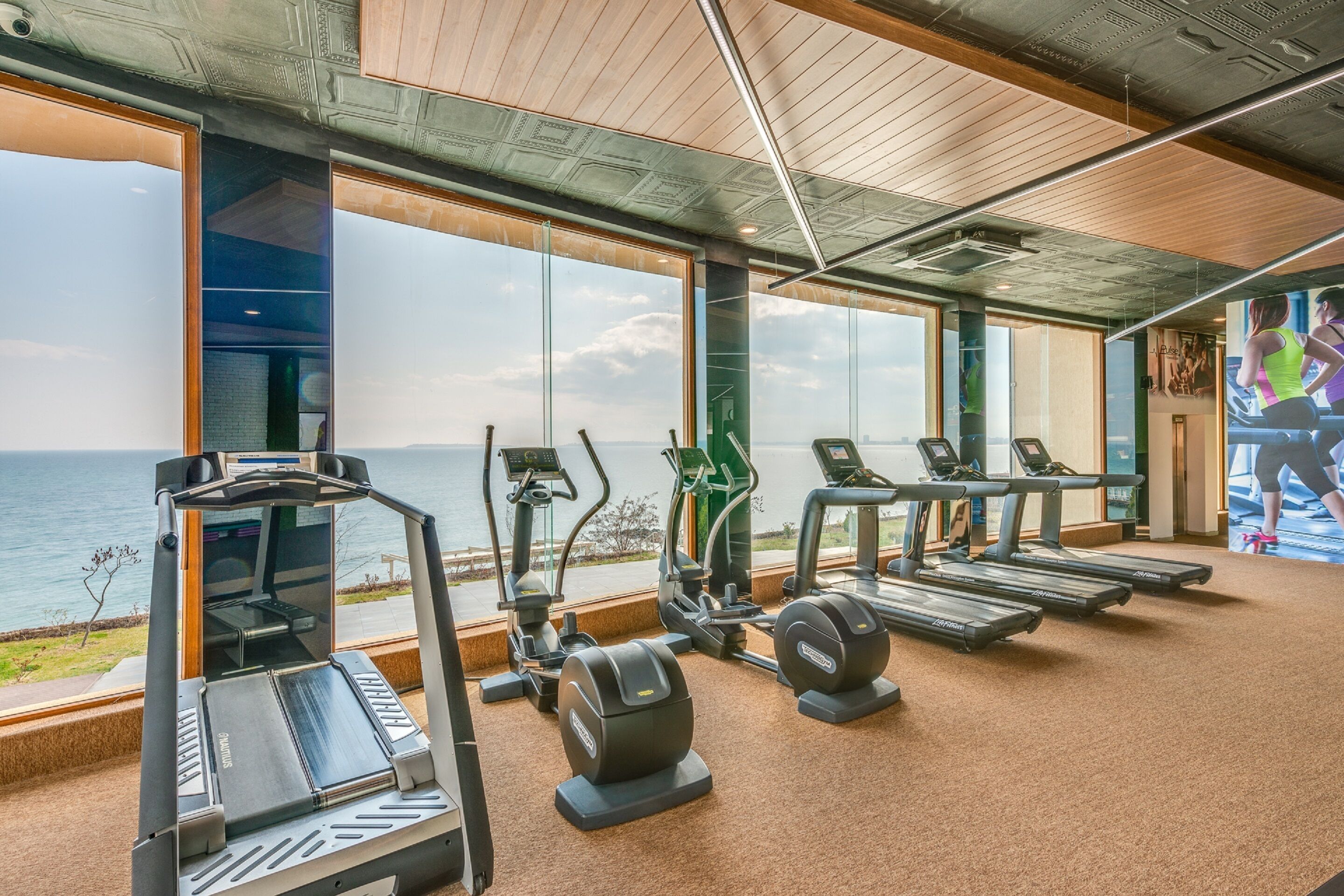 A gym with treadmills, elliptical machines, and stationary bikes, large windows with a view of the sea, and a high ceiling with exposed beams.