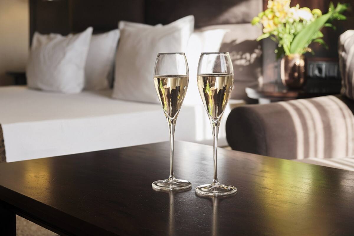 Två champagneglas på ett träbord i ett hotellrum med en säng och ett nattduksbord med en vas med blommor.