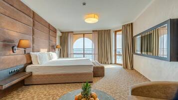 Habitación de hotel con una cama grande, un televisor de pantalla plana, una mesa de centro redonda con fruta y un balcón con cortinas.