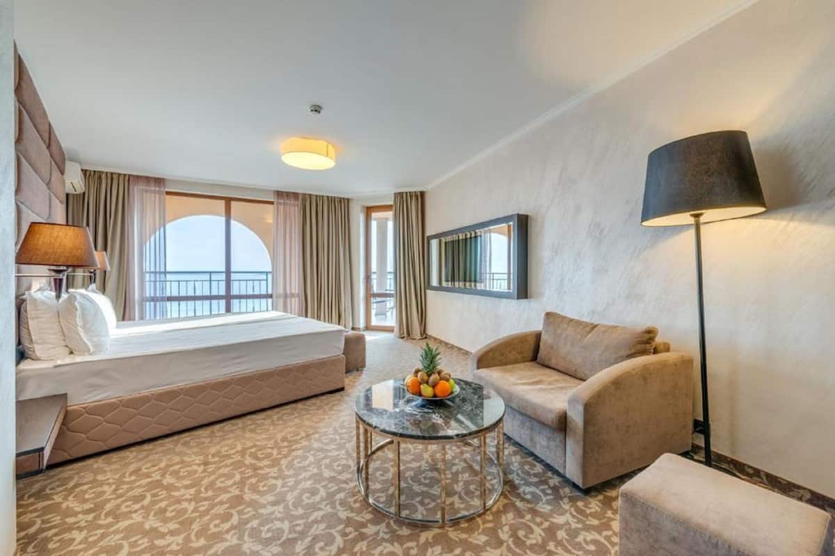 Ett hotellrum med en säng, en soffa, ett runt bord med frukt på och ett stort fönster med gardiner.