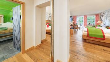 Ein Hotelzimmer mit Holzboden, einem Bett mit grün-roter Bettwäsche, einem Schreibtisch mit Stuhl und Blick auf Bäume durch das Fenster.
