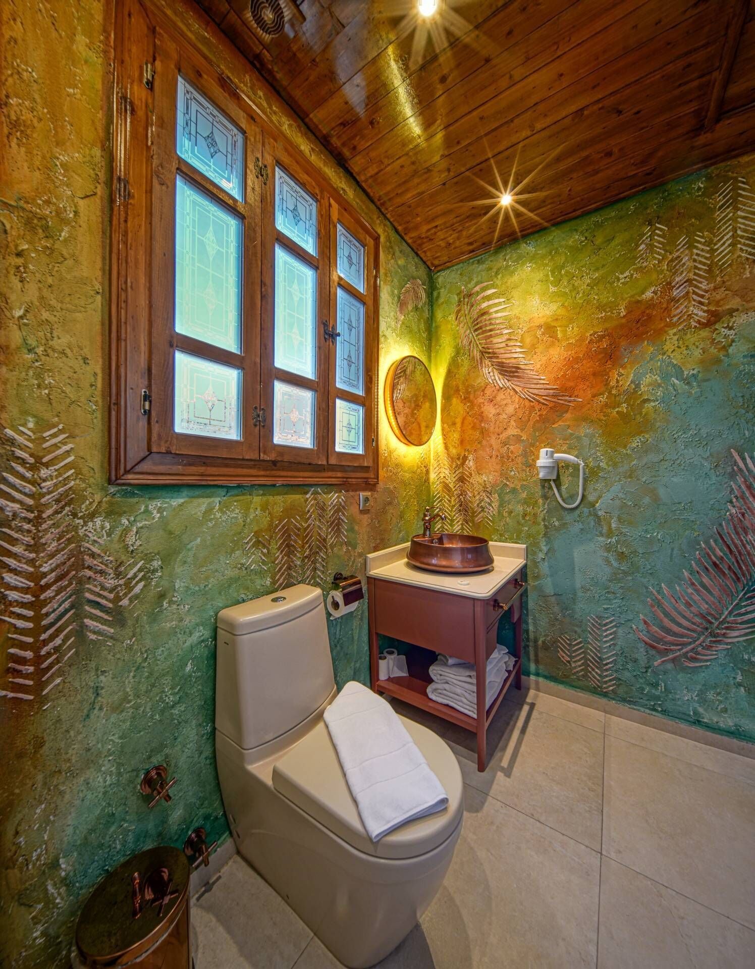 A bathroom with a wooden ceiling, a toilet, a sink with a copper basin, and a window with stained glass.