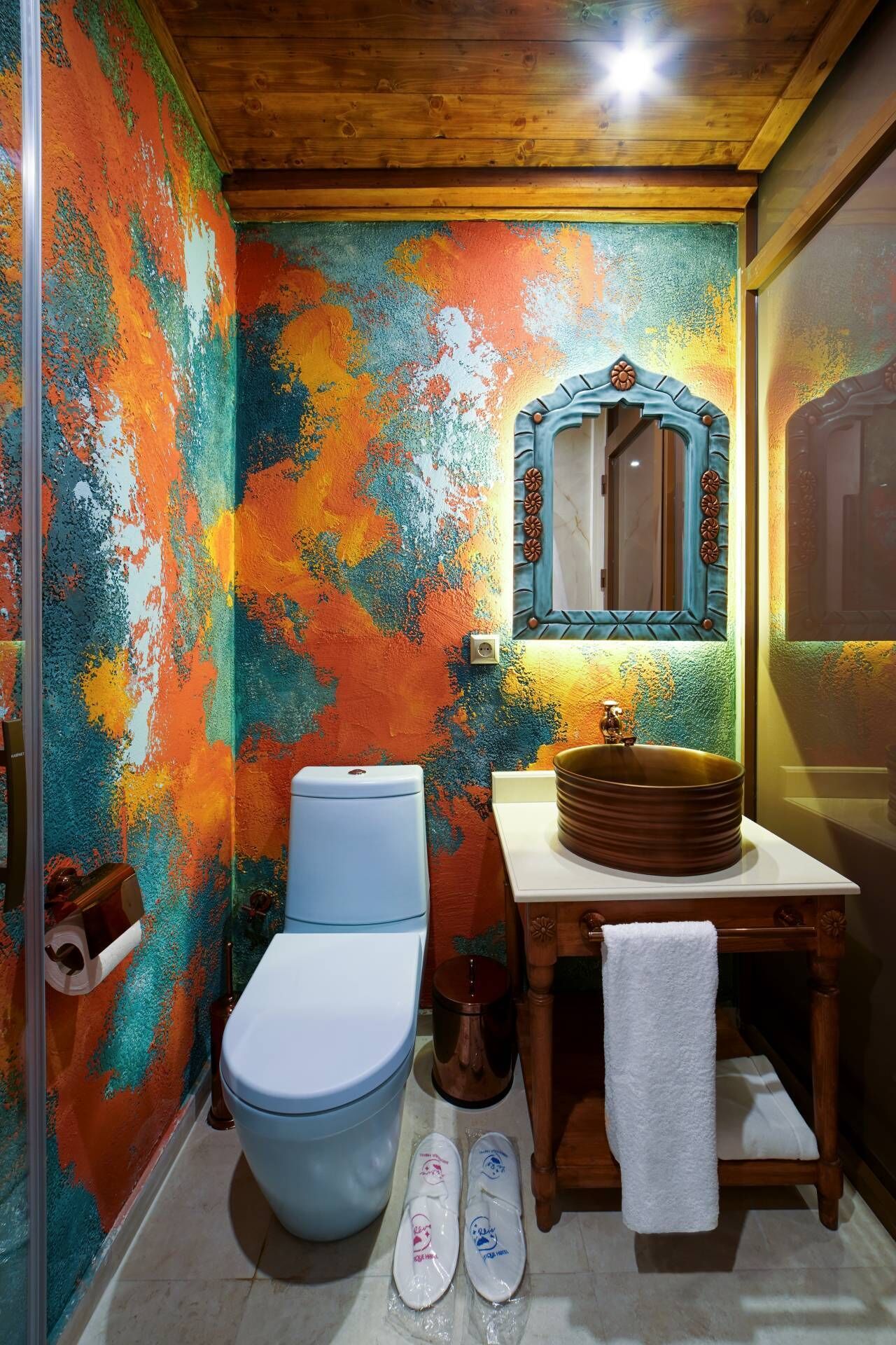 Un baño con una pared de diseño abstracto y colorido, un inodoro blanco, un tocador de madera con un lavabo redondo y un espejo con marco decorativo.