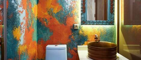 Un baño con una pared de diseño abstracto y colorido, un inodoro blanco, un tocador de madera con un lavabo redondo y un espejo con marco decorativo.