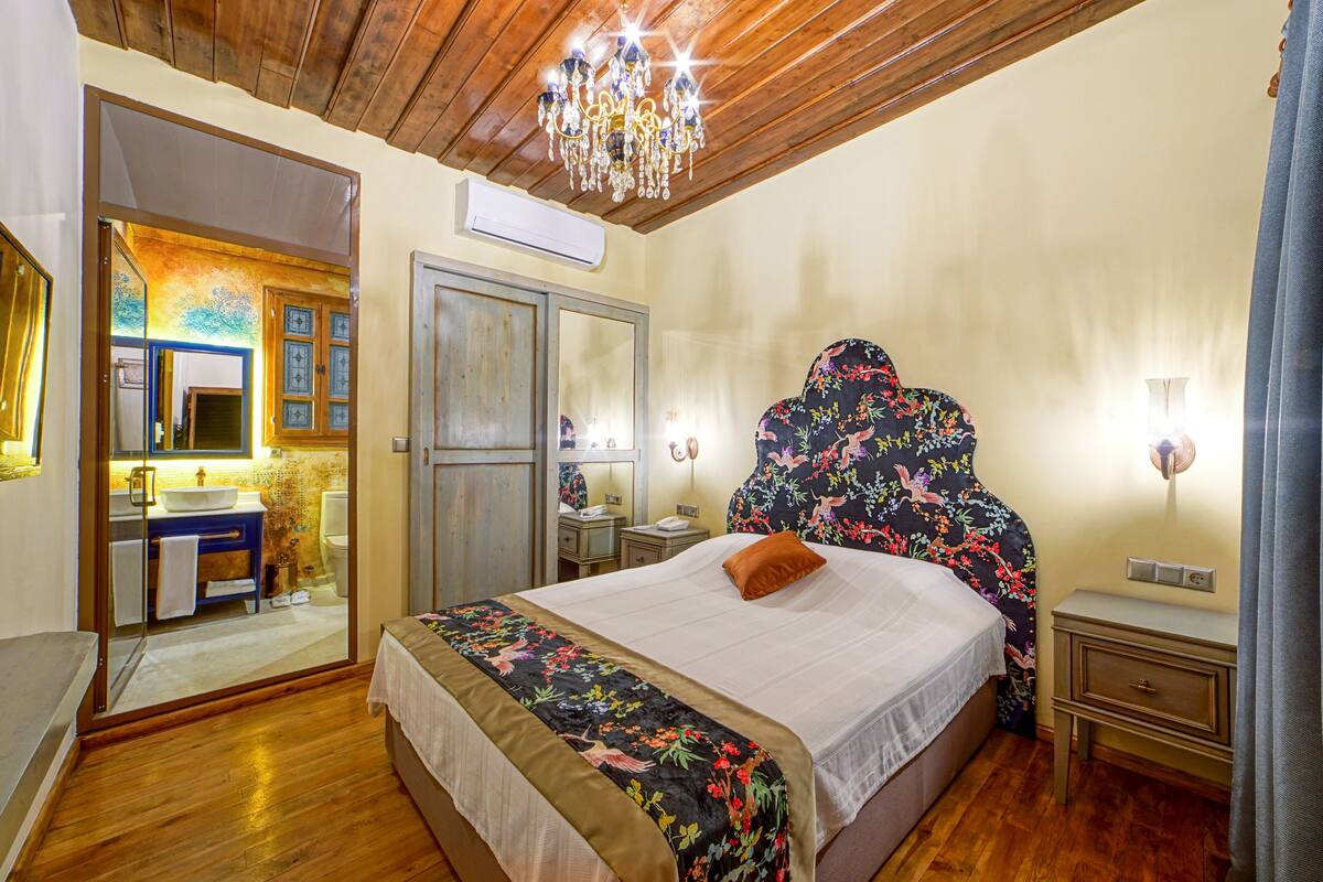 Un dormitorio con techo de madera, una araña, una cama con cabecera floral, una mesita de noche, un aparador y un baño visible a través de una puerta de vidrio.