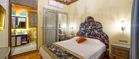 Un dormitorio con techo de madera, una araña, una cama con cabecera floral, una mesita de noche, un aparador y un baño visible a través de una puerta de vidrio.