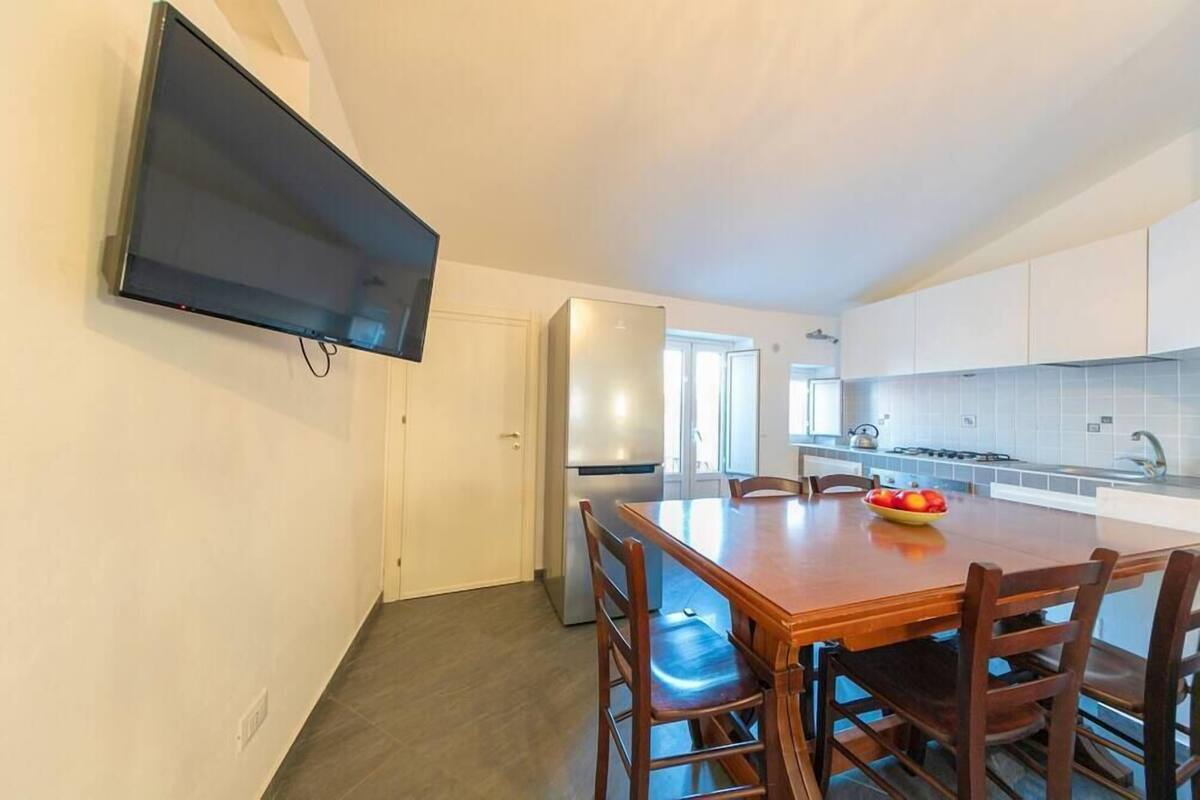 A modern kitchen with white cabinets, a stainless steel refrigerator, a wooden dining table with chairs, and a flat-screen TV mounted on the wall.