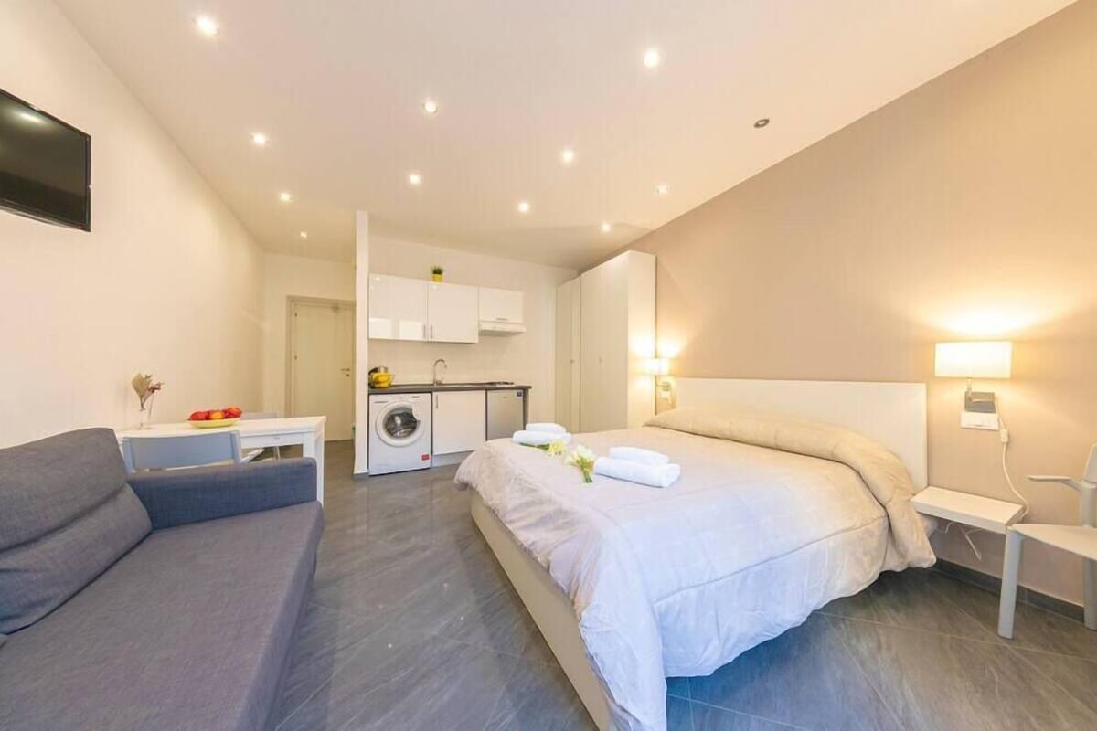 A modern hotel room with a large bed, a sofa, a small table, and a kitchenette in the background.