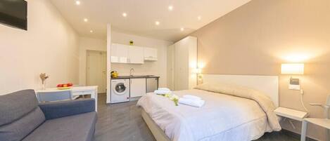 A modern hotel room with a large bed, a sofa, a small table, and a kitchenette in the background.