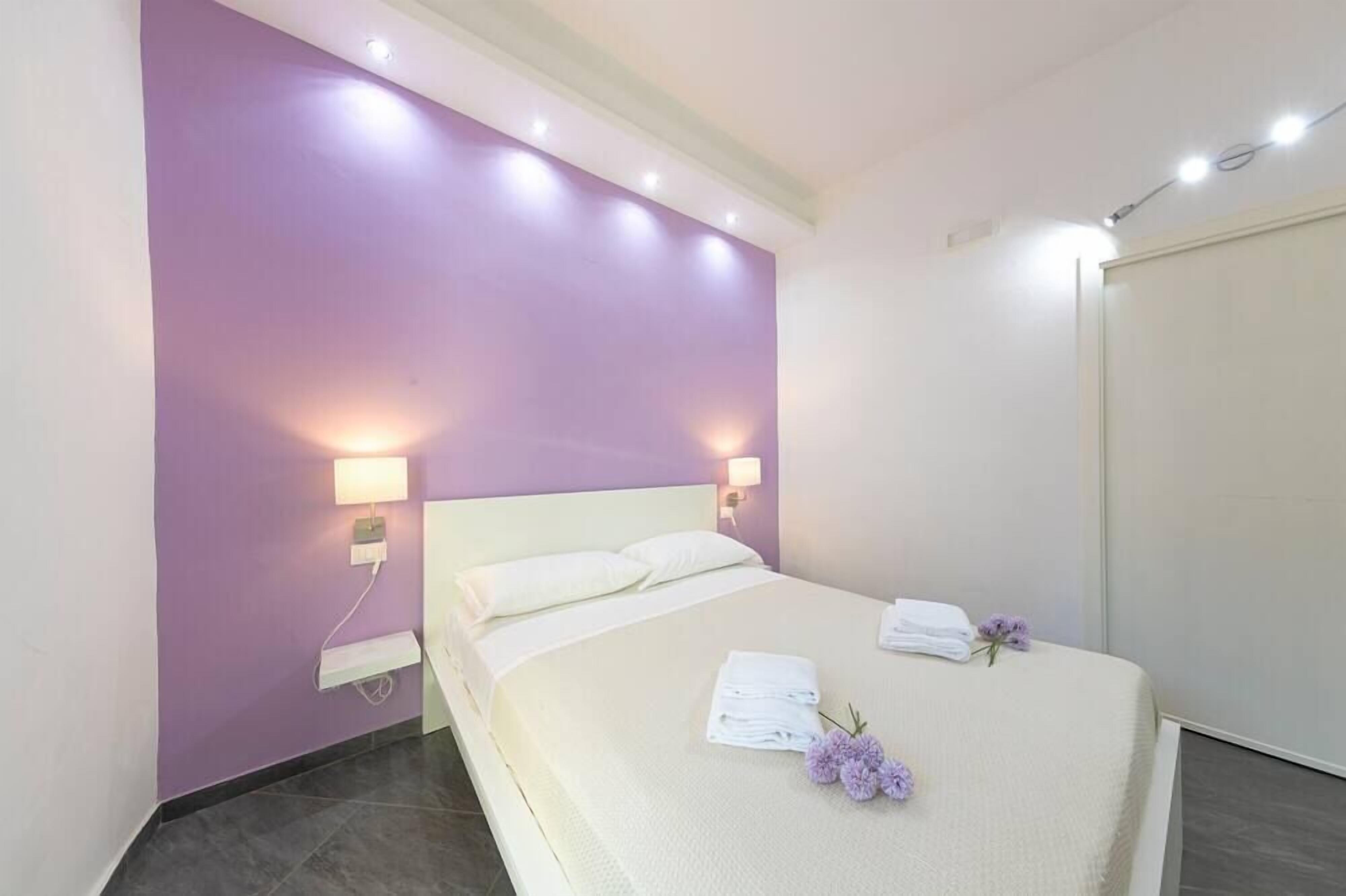 Een slaapkamer met een paarse wand, een bed met witte beddengoed, twee bedlampen en een nachtkastje.
