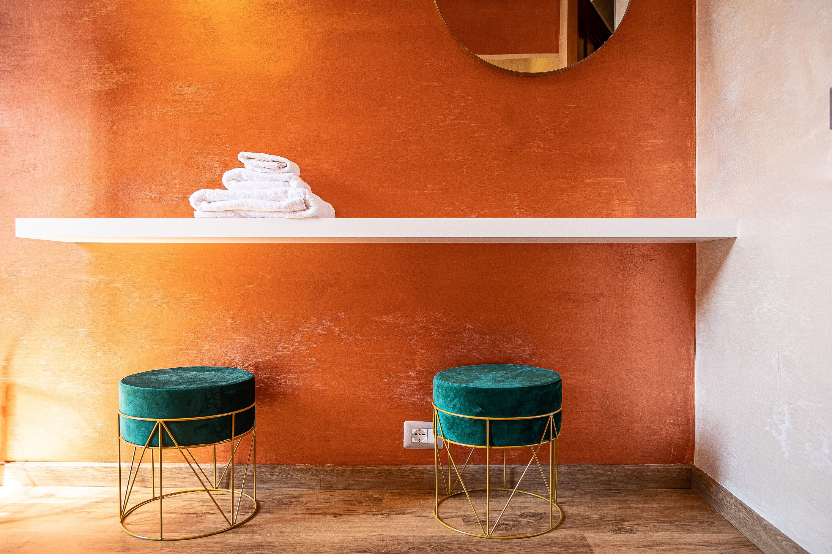 Een hoek met een witte plank met gevouwen handdoeken, twee groene fluweelstoelen met gouden draadframes en een houten vloer.