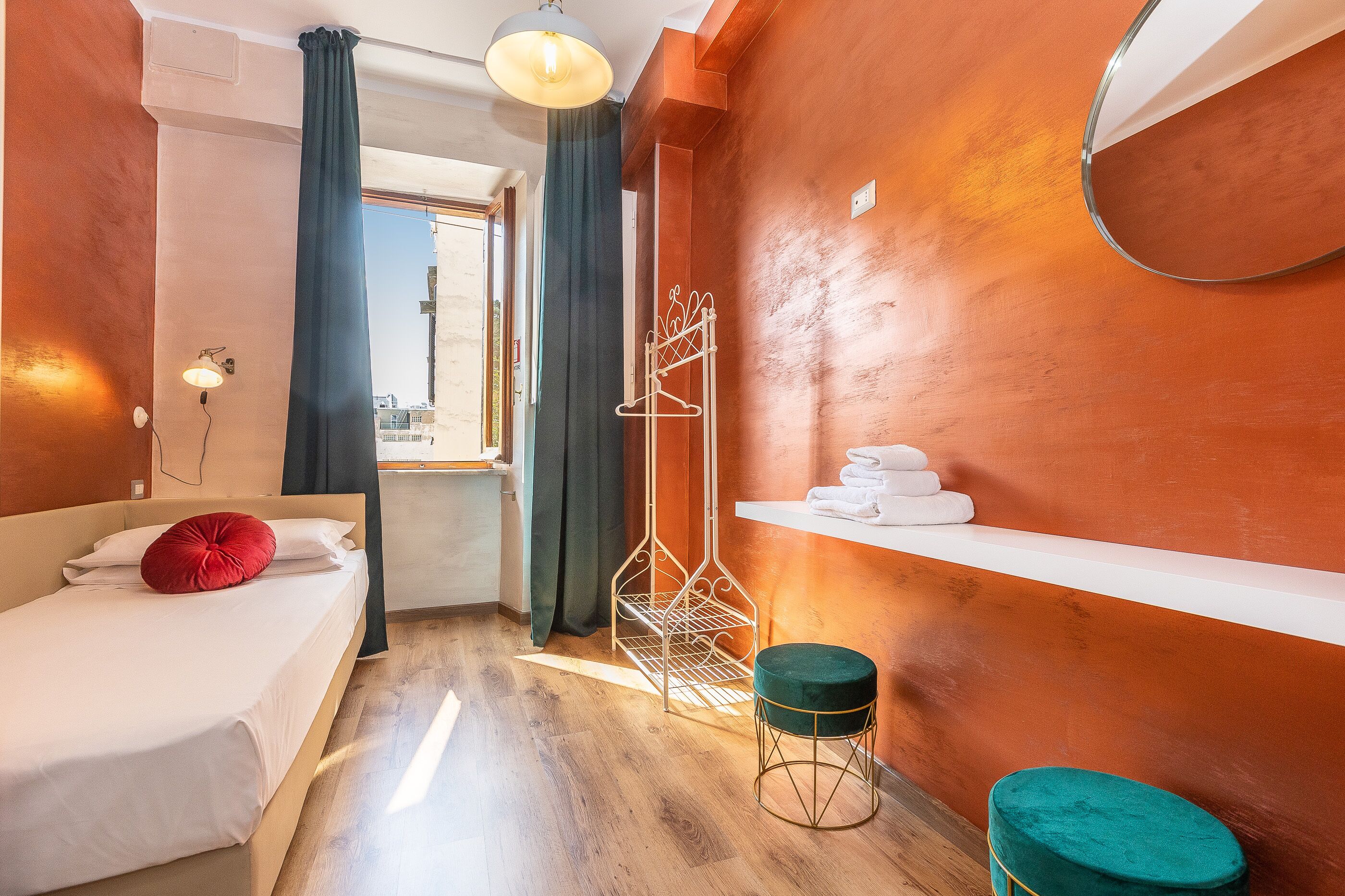 A hotel room with a bed, a small stool, a metal rack, and a shelf with towels.