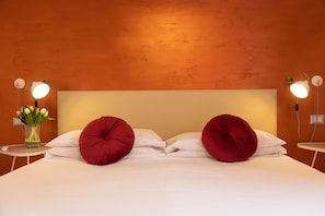 A neatly made bed with two large red pillows, a white bedside table with a vase of flowers, and two wall-mounted lamps. - Lovely New Home, Via Genova (Roma)