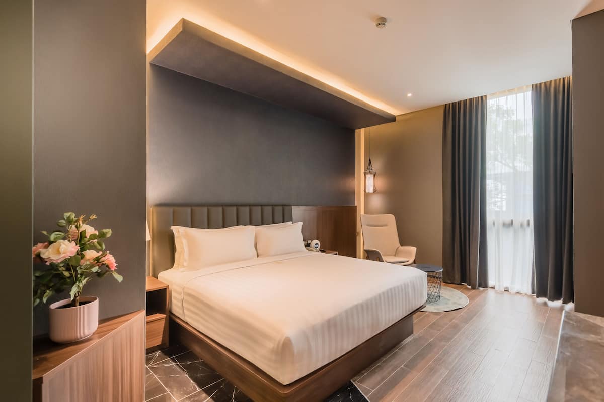 Une chambre d’hôtel moderne avec un grand lit, des tables de chevet, une plante en pot, une chaise et une vue sur l’extérieur par une fenêtre.