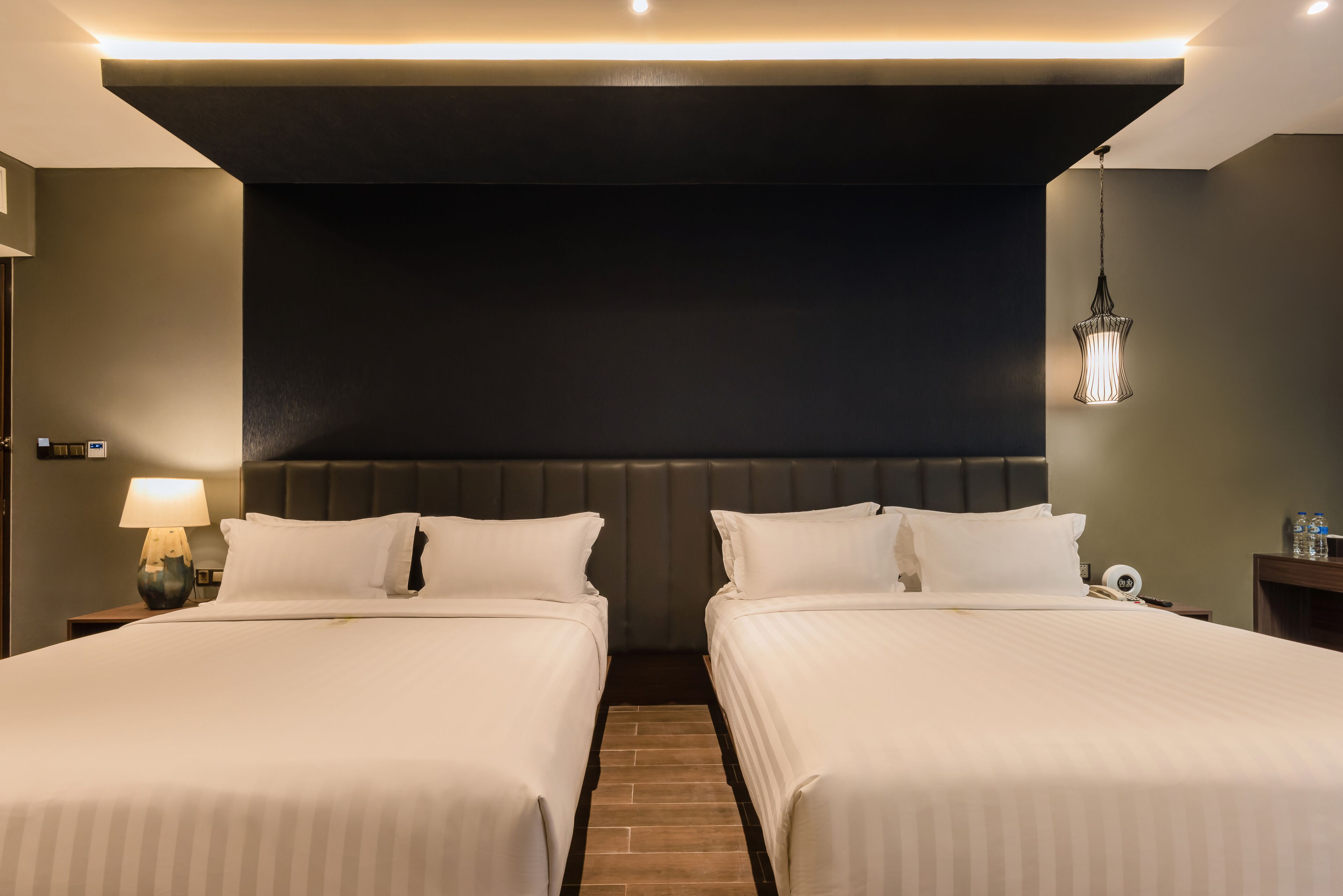 A hotel room with two beds, a dark headboard, a nightstand with a lamp, and a hanging light fixture.