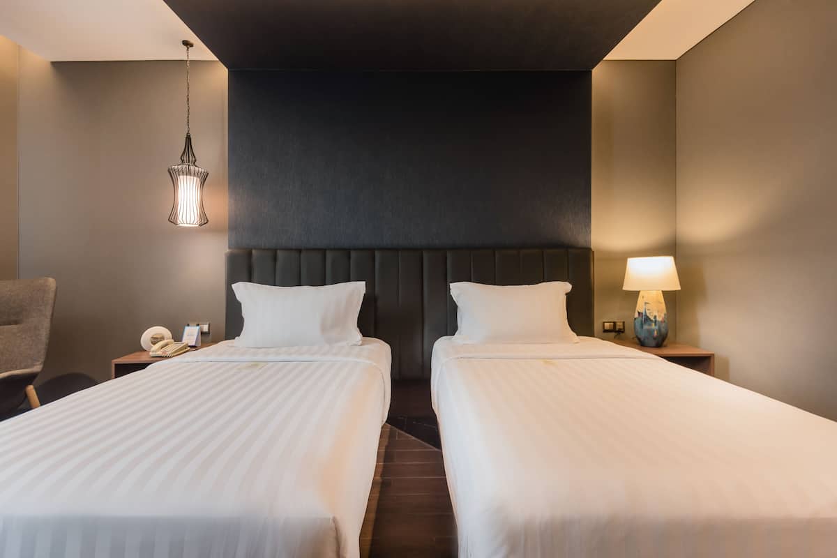 Une chambre d’hôtel avec deux lits, une tête de lit, un luminaire suspendu, une chaise, une table de chevet avec une lampe et un téléphone.