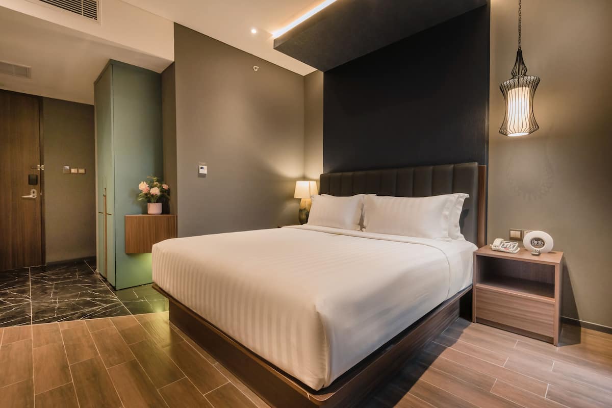 Une chambre d’hôtel moderne avec un grand lit, une table de chevet avec une lampe, une petite table de nuit avec un téléphone et une horloge, et un vase décoratif avec des fleurs.