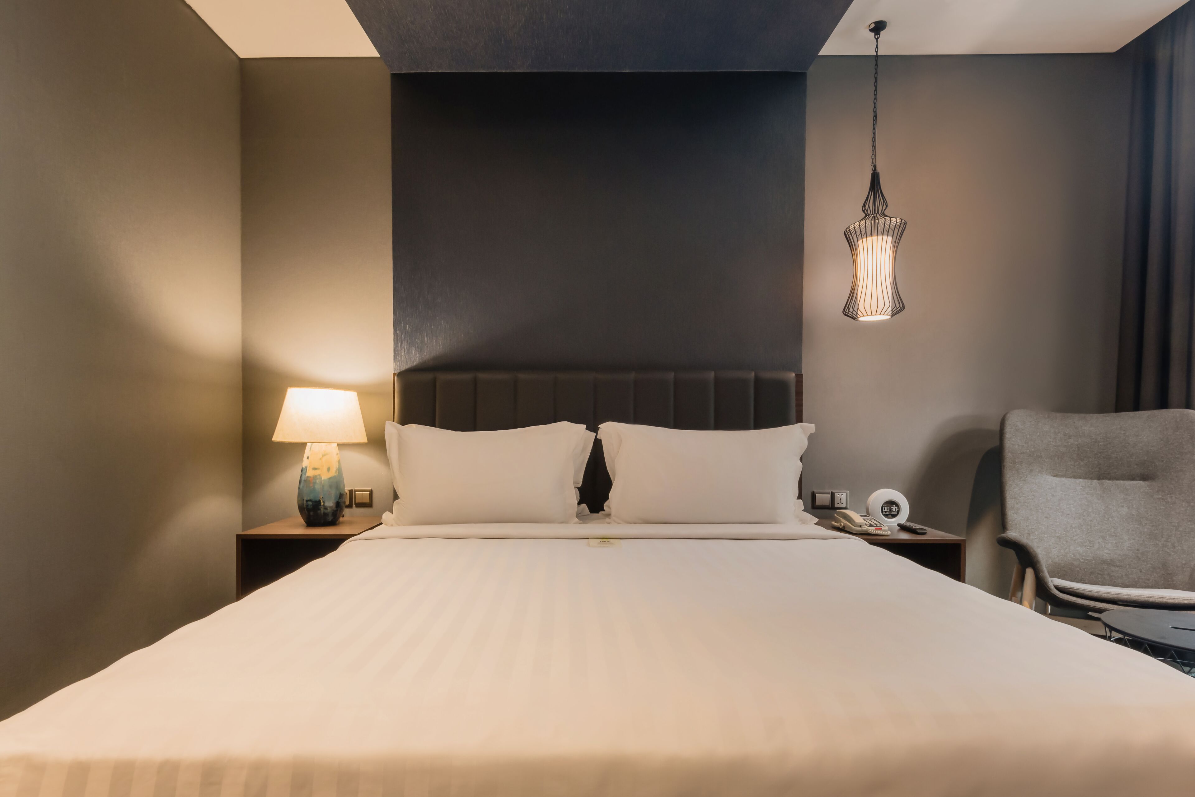 A hotel room with a large bed, a nightstand with a lamp, a chair, and a hanging light fixture.