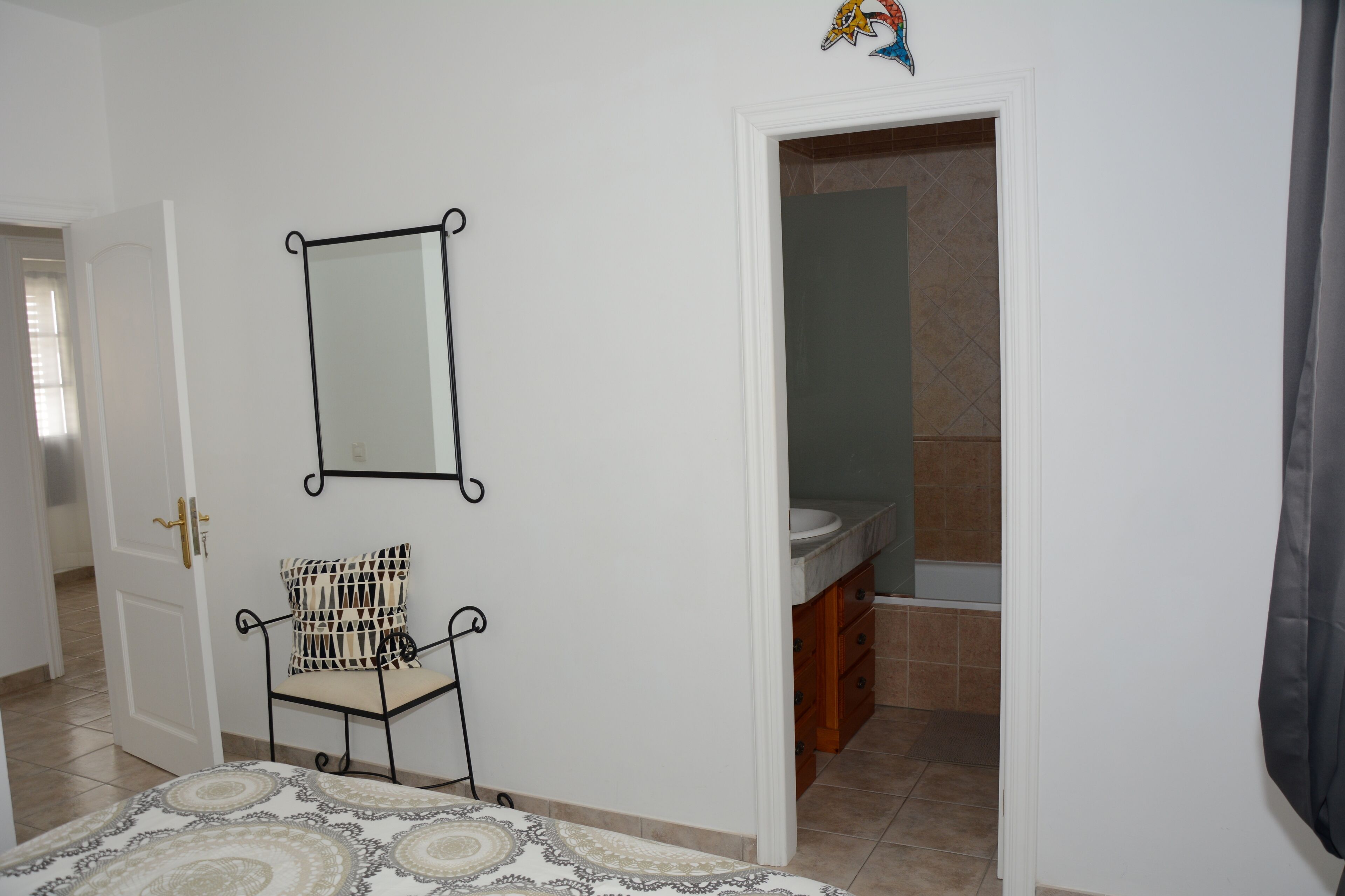 A bedroom with a bed, a chair, a mirror, and a bathroom visible through an open door.
